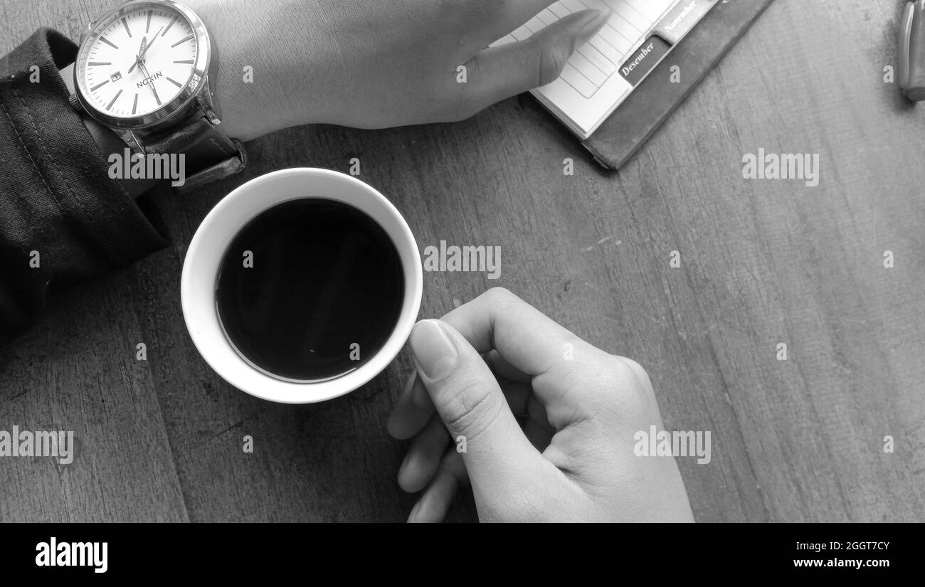 WONOSOBO, INDONESIA - Apr 07, 2021: A grayscale shot of a person drinking black coffee and browsing through a notebook Stock Photo