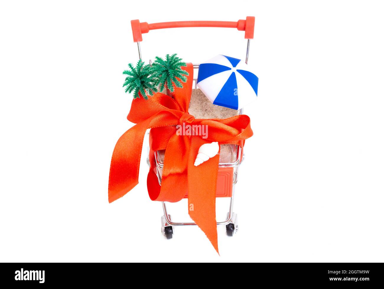 Tropical beach set-up in a miniature shopping cart with a big red bow isolated on white. Gifting a beach vacation. Stock Photo