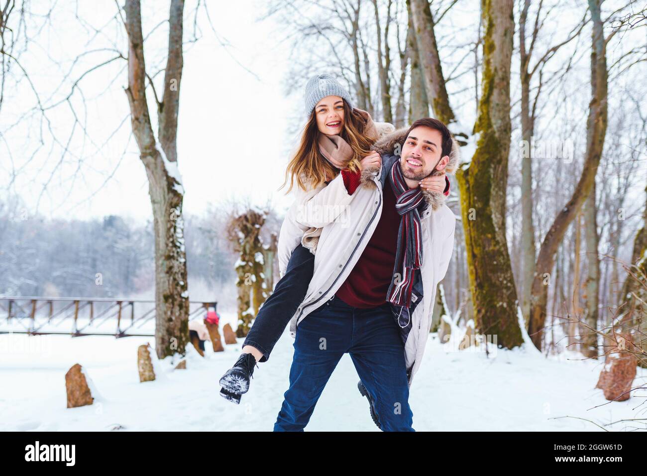 Happy young man carry his smiling pretty woman on his back at winter park. Boyfriend giving piggyback ride on shoulder to his girlfriend in winter hol Stock Photo