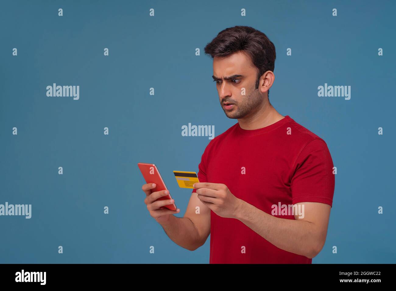 Portrait of a young man holding a credit card looking at his phone with a shocked expression. Stock Photo