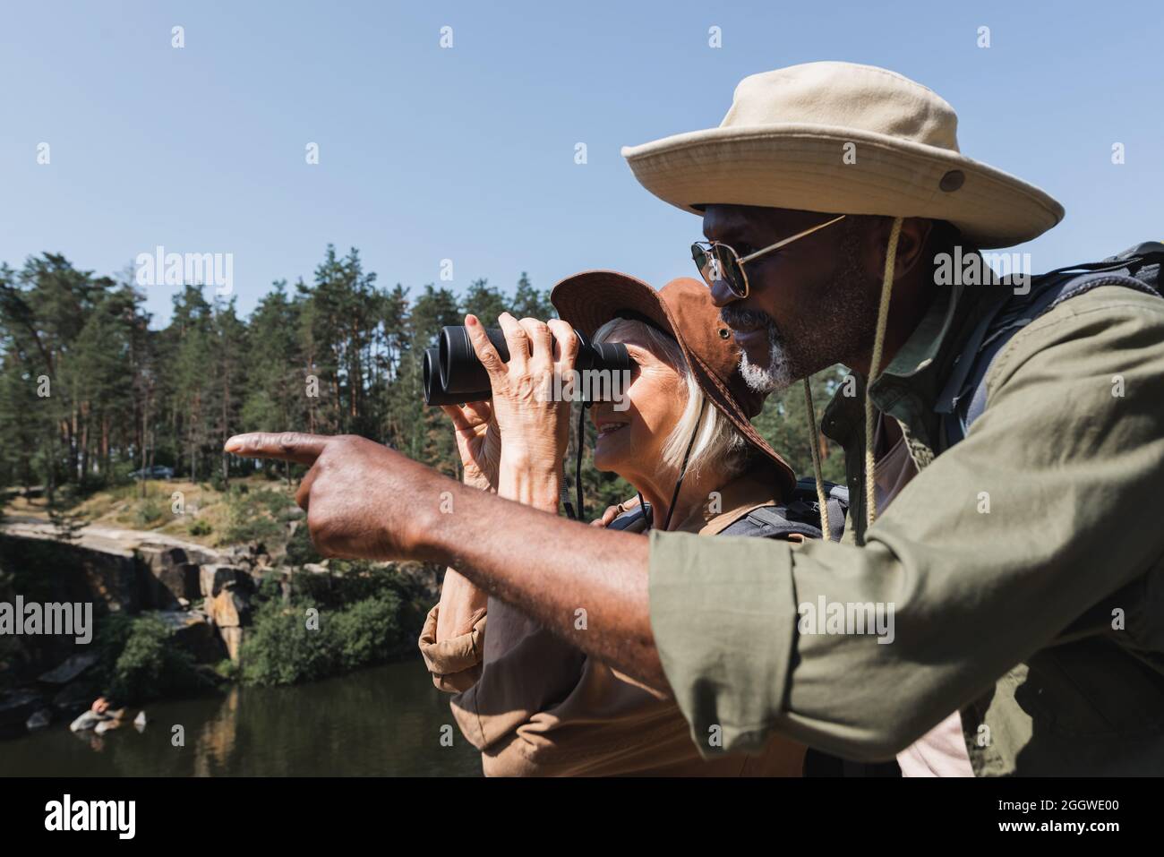 Side view of african american hiker pointing with finger near smiling wife with binoculars outdoors Stock Photo