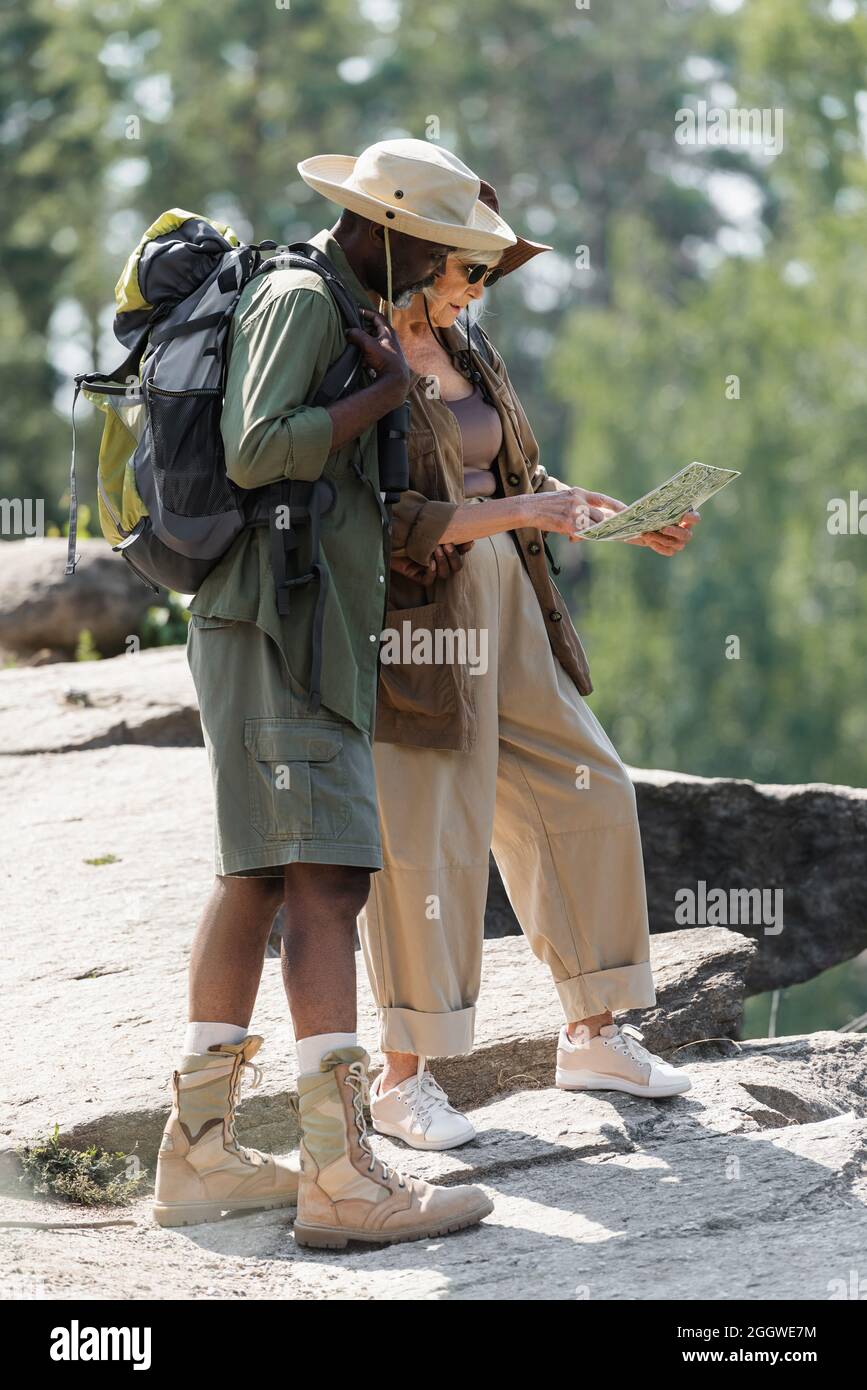 Side view of senior hiker pointing at map near african american husband on rock Stock Photo