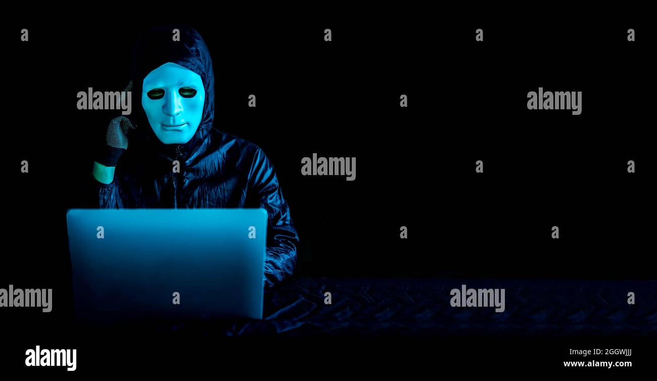 Hacker in mask and hoodie using a laptop to browse the net and serious face thinking about question Stock Photo