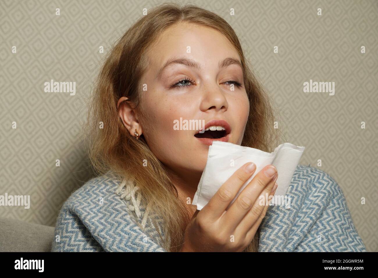 Woman sneezes in white tissue.  Young blonde woman wrapped in warm blanket, sneezes, has flu. Stock Photo