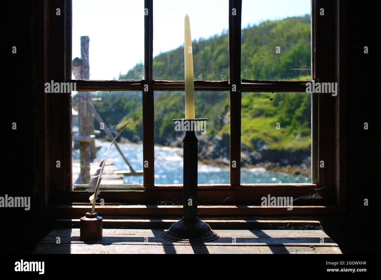 White candle in an old fashioned candle holder, on top of an old desk, next to a quill pen in an ink well, in front of a window, overlooking the sea. Stock Photo