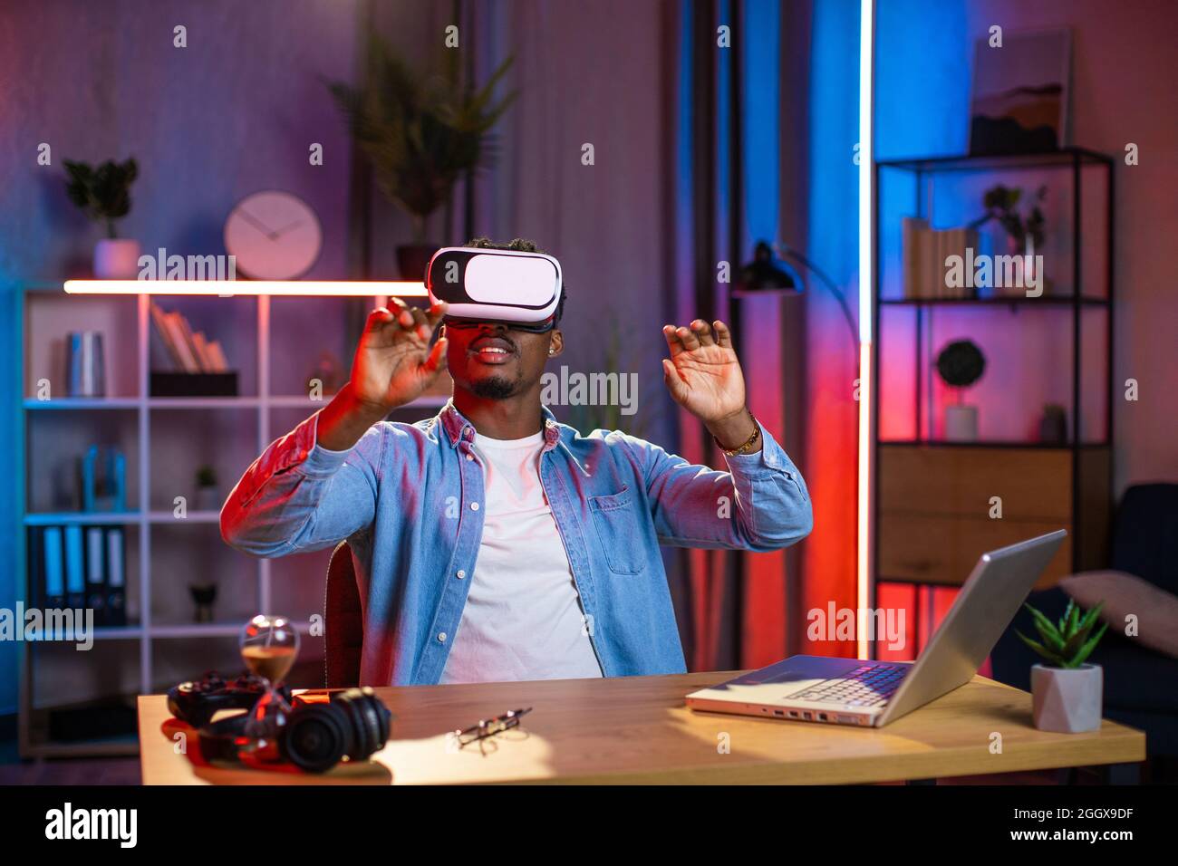 Young black guy in VR goggles working in augmented reality while staying at  home. Handsome man in denim shirt sitting at desk with laptop and tapping  on virtual screen Stock Photo -