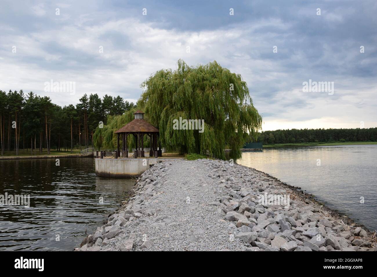 Gazebo under a large tree on the shore of a lake with a rocky embankment and a sky with clouds Stock Photo