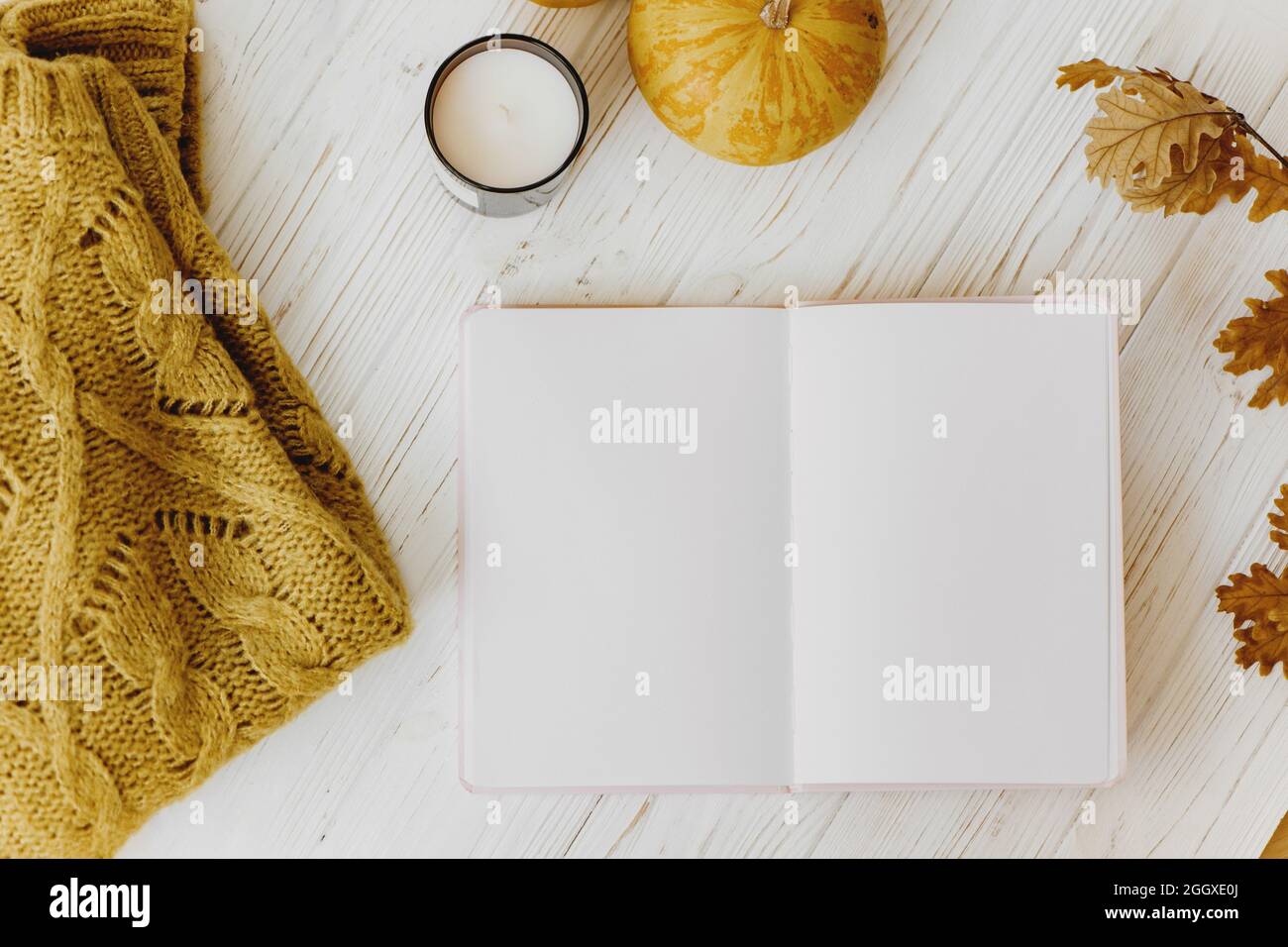 Autumn flat lay with opened notebook, warm sweater, orange pumpkin, candle and yellow autumn leaves on white wooden background. Stock Photo