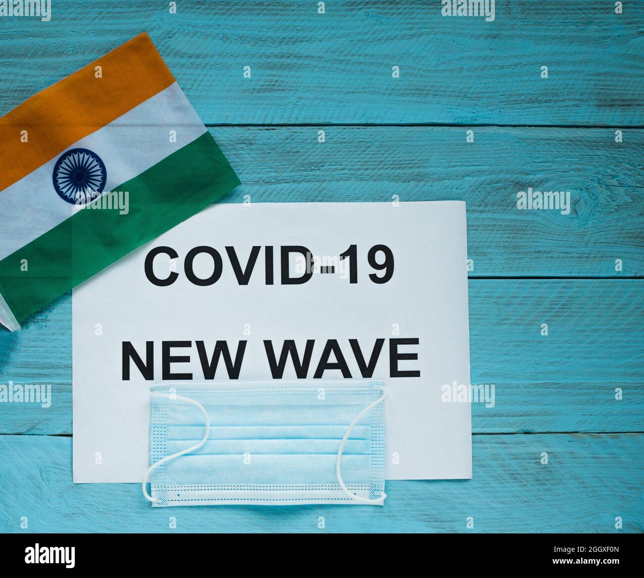 On a blue wooden background lie a sheet with the inscription: covid -19 new wave, a medical mask and a flag of India. Concept of the next wave of covi Stock Photo