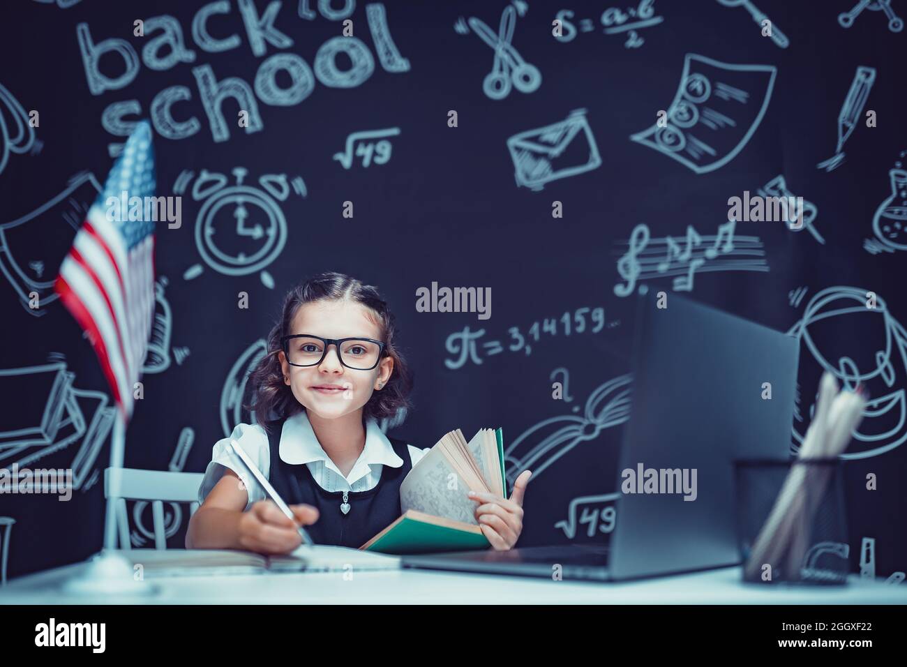 beautiful little schoolgirl sitting at desk and study online with laptop against black background with USA flag Stock Photo