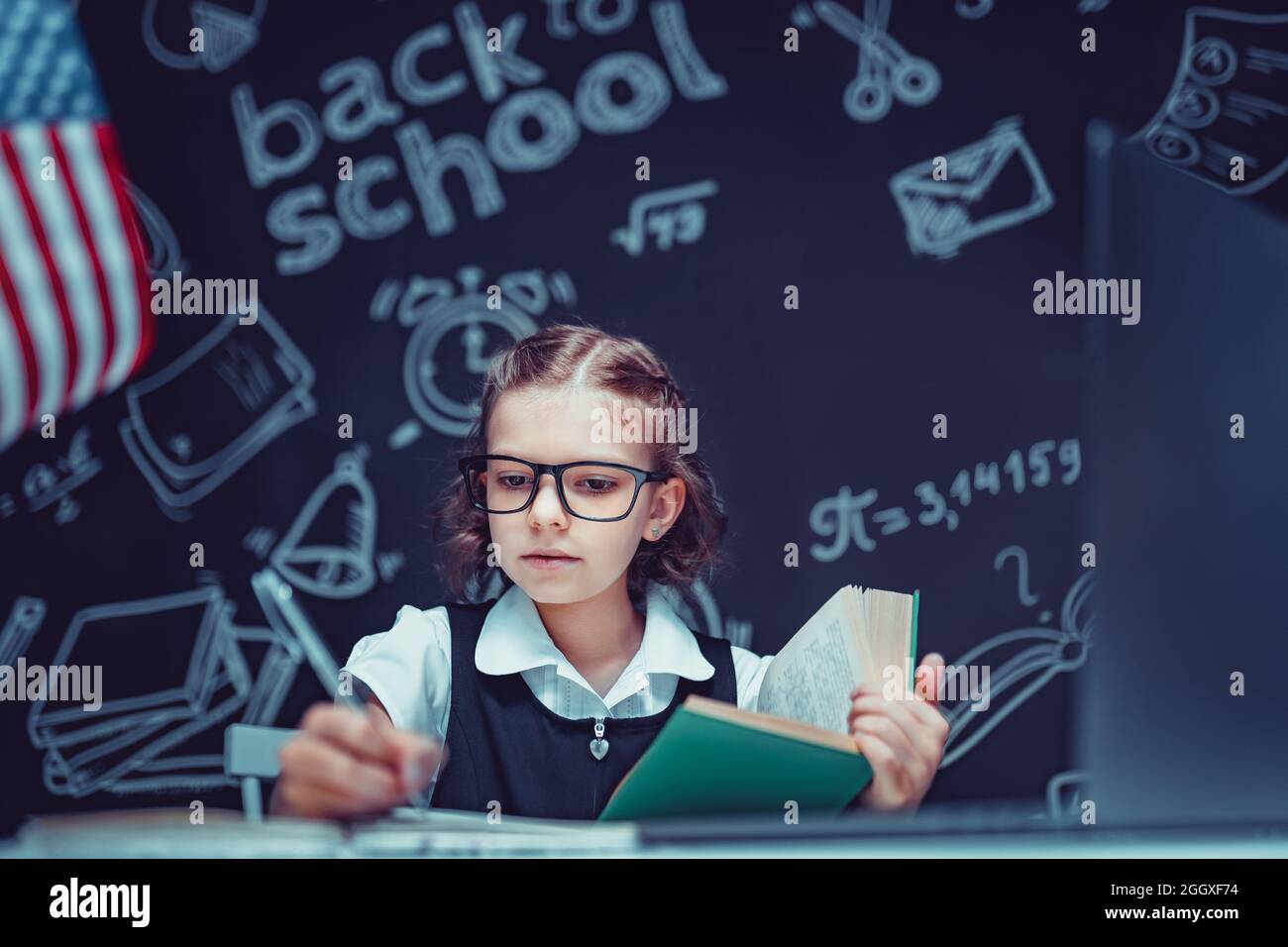 beautiful little schoolgirl sitting at desk and study online with laptop against black background with USA flag Stock Photo