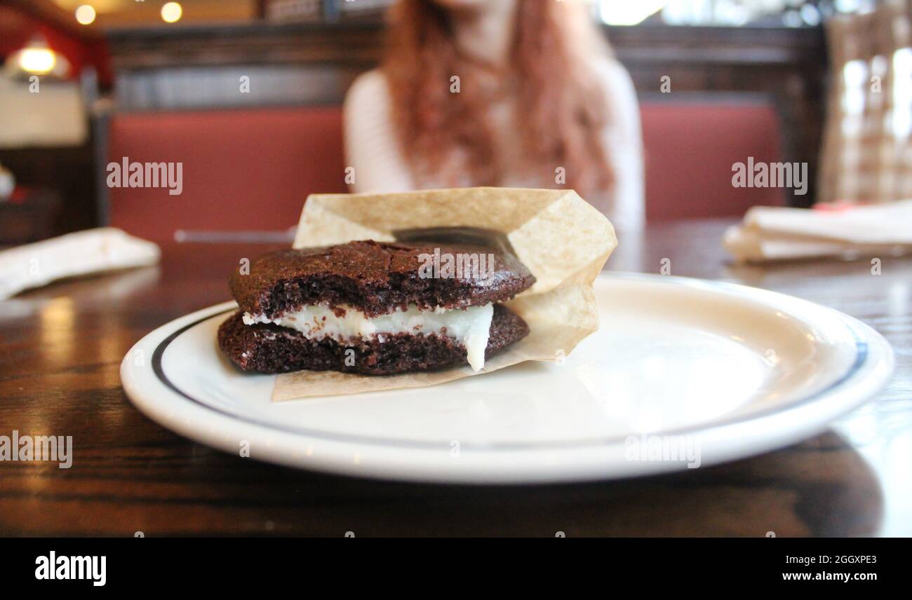 a whoppie pie, sometimes spelled whoppy, a classic sweet pastry with a bite taken, sitting on a plate inside a sit down restaurant with a woman Stock Photo
