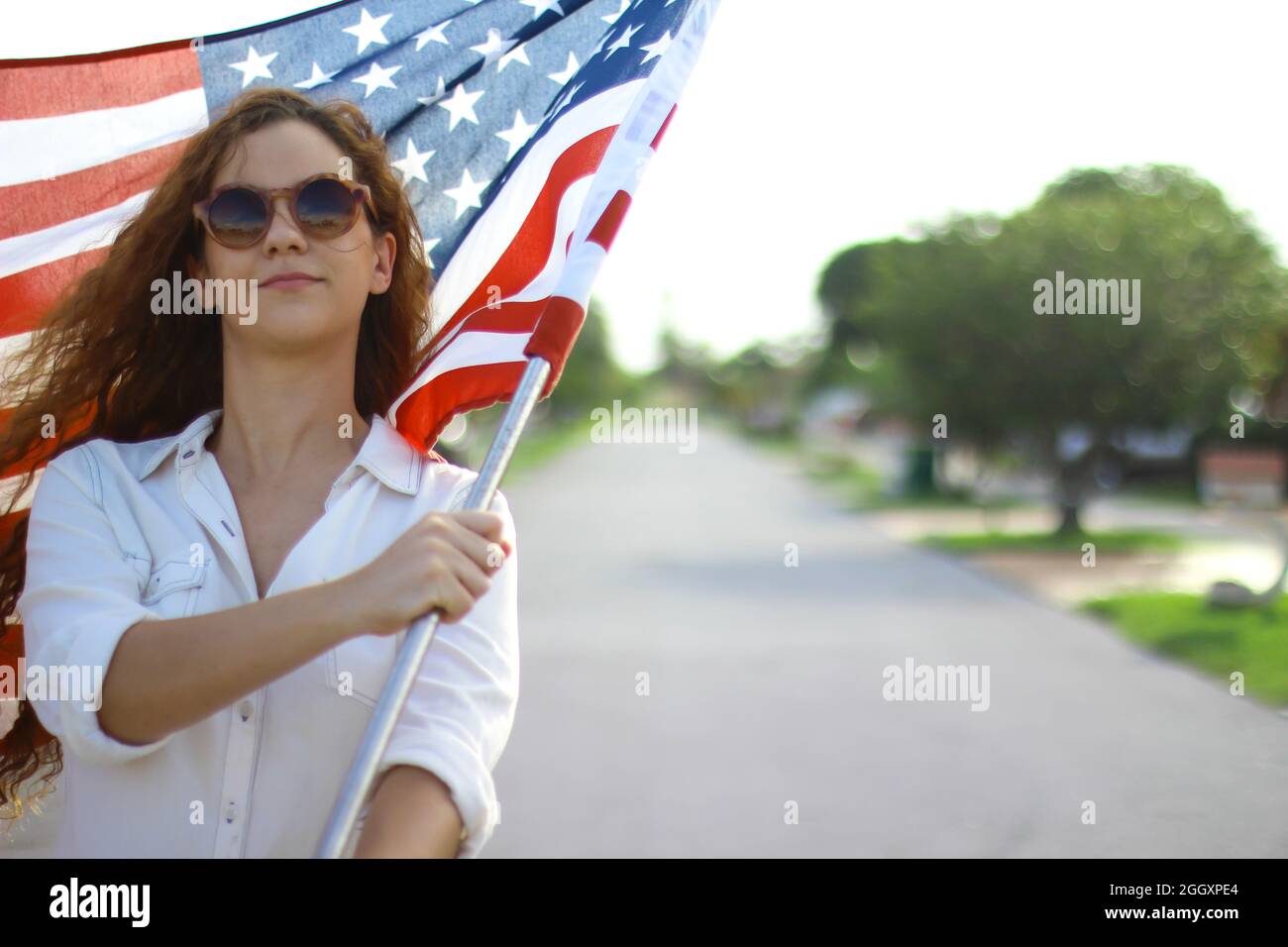 Young redhead Hispanic and Caucasian woman wearing sunglasses waving the American flag outside. Stock Photo