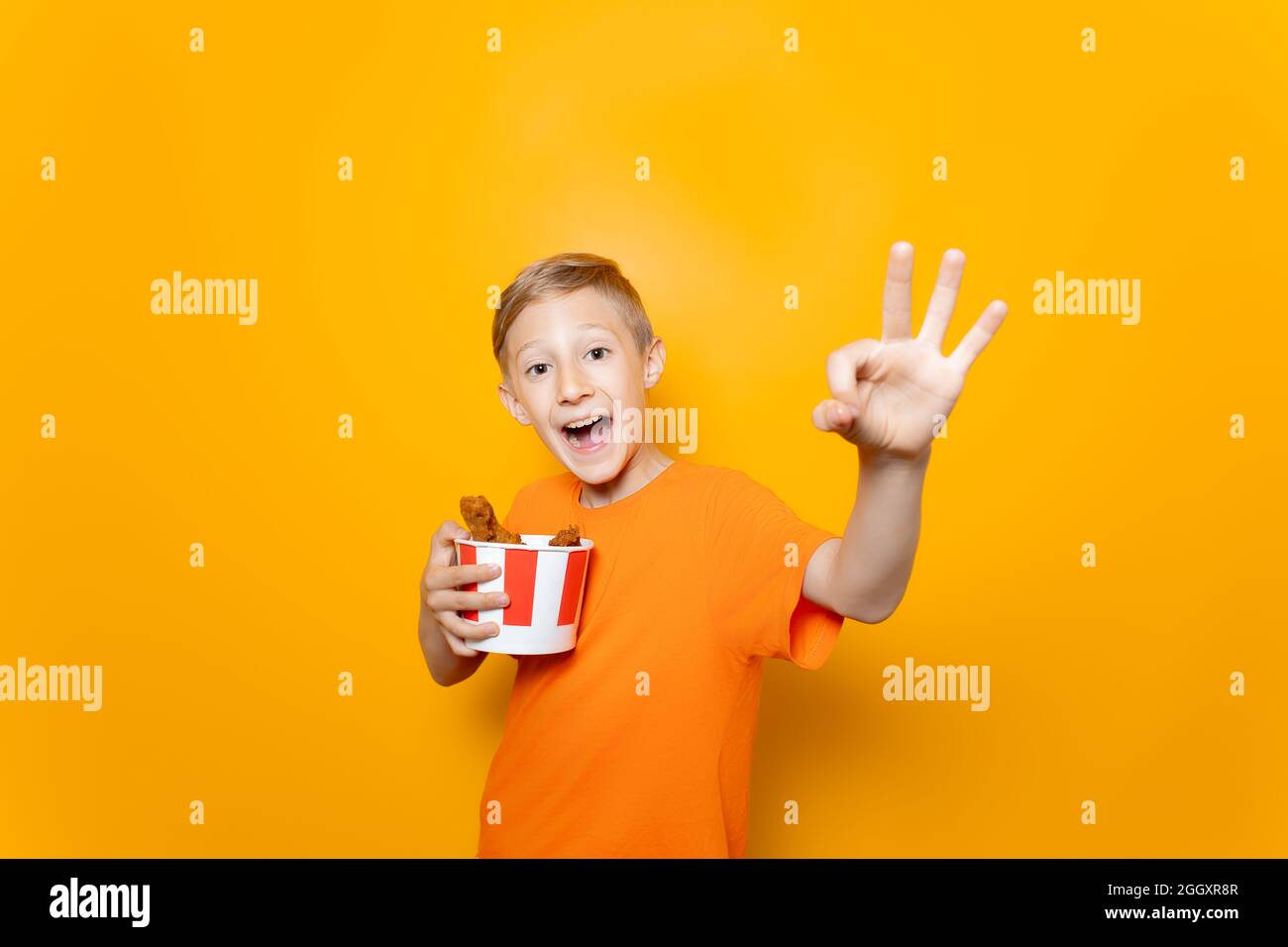 a boy in an orange T-shirt holds a paper bucket of chicken wings in front of him and shows the Ok sign Stock Photo