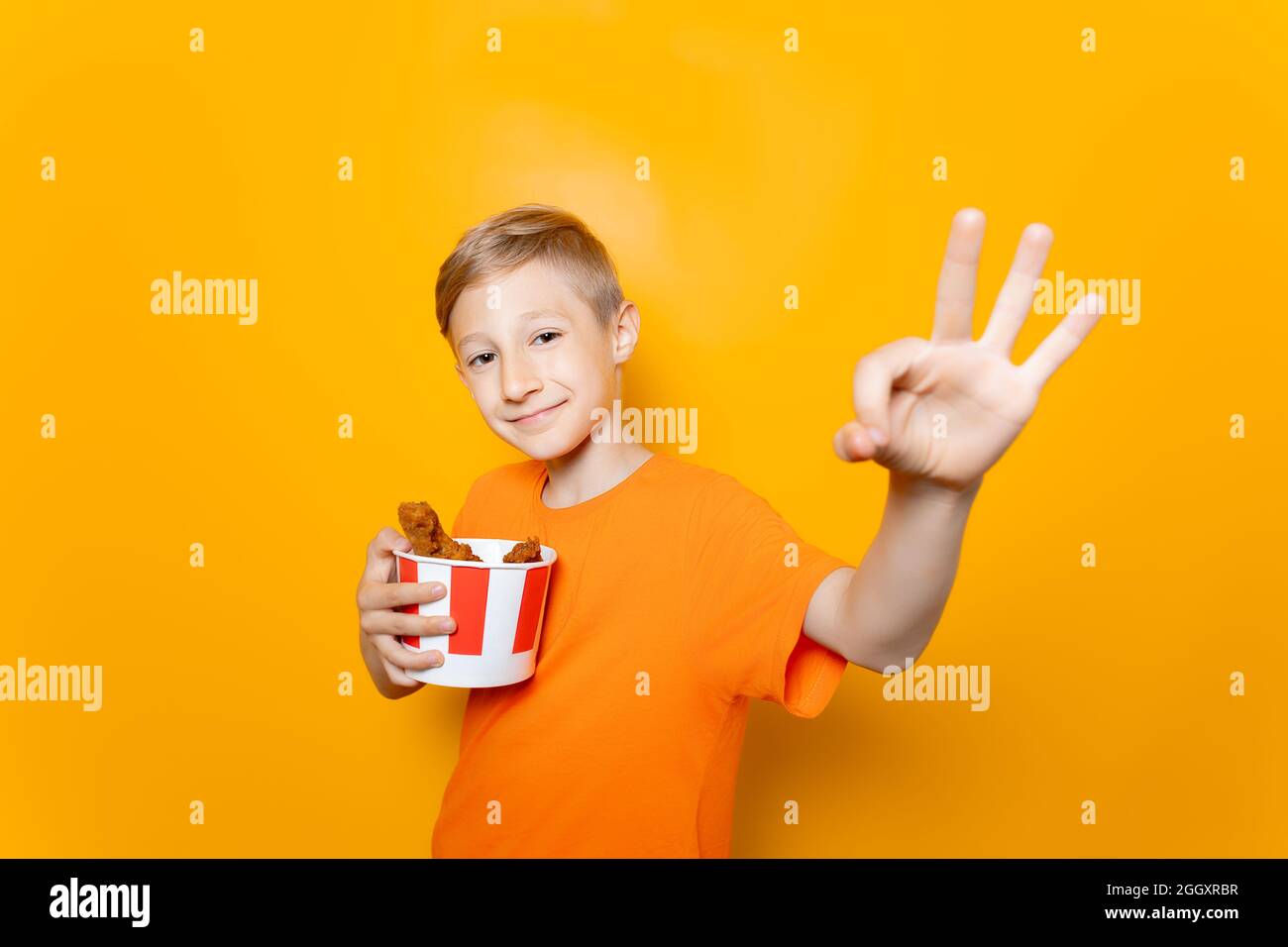 a boy in an orange T-shirt holds a paper bucket of chicken wings in front of him and shows the Ok sign Stock Photo