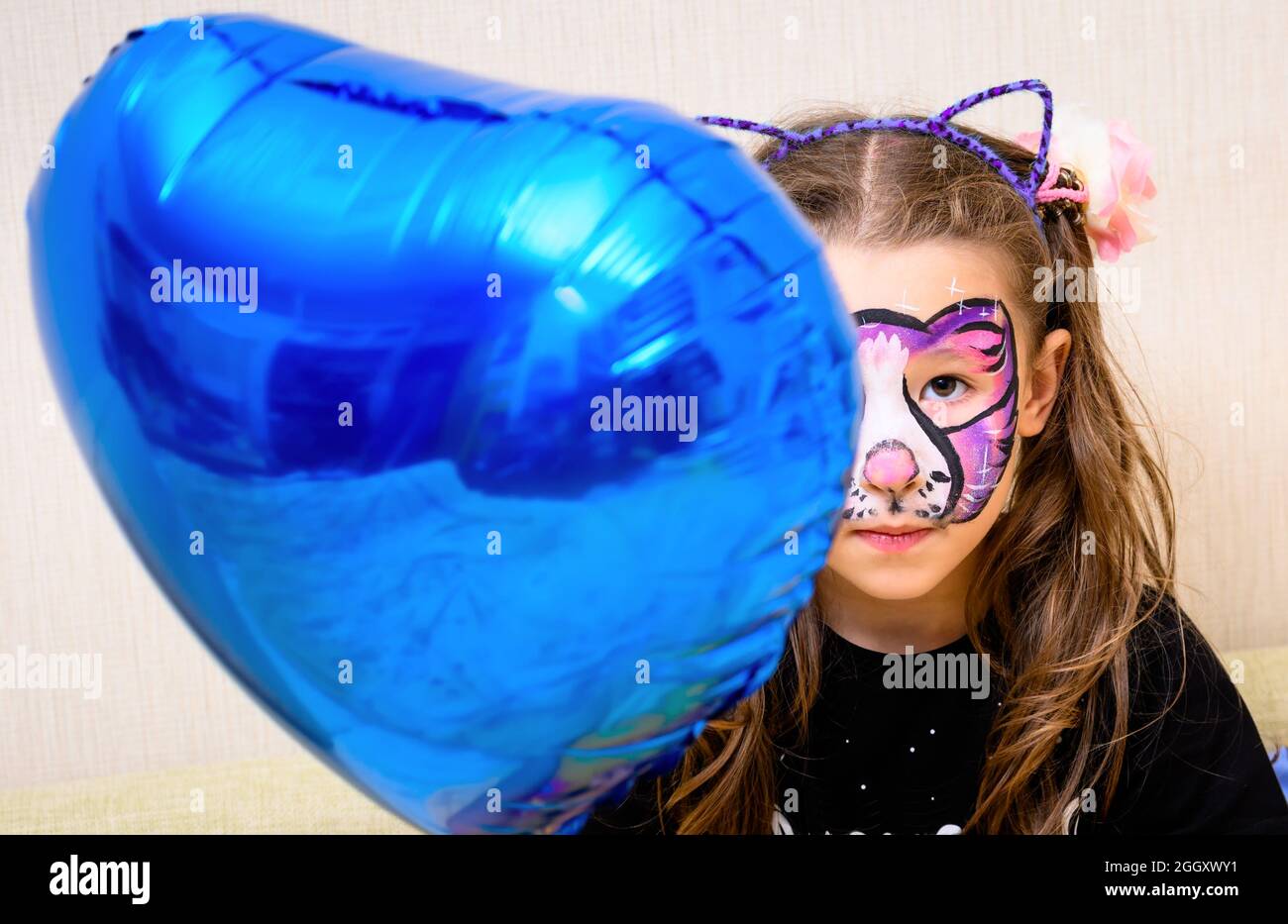Kid with face painting of kitty holds foil balloon, cute little girl with painted colorful mask of cat on her face. Portrait of serious child with bea Stock Photo