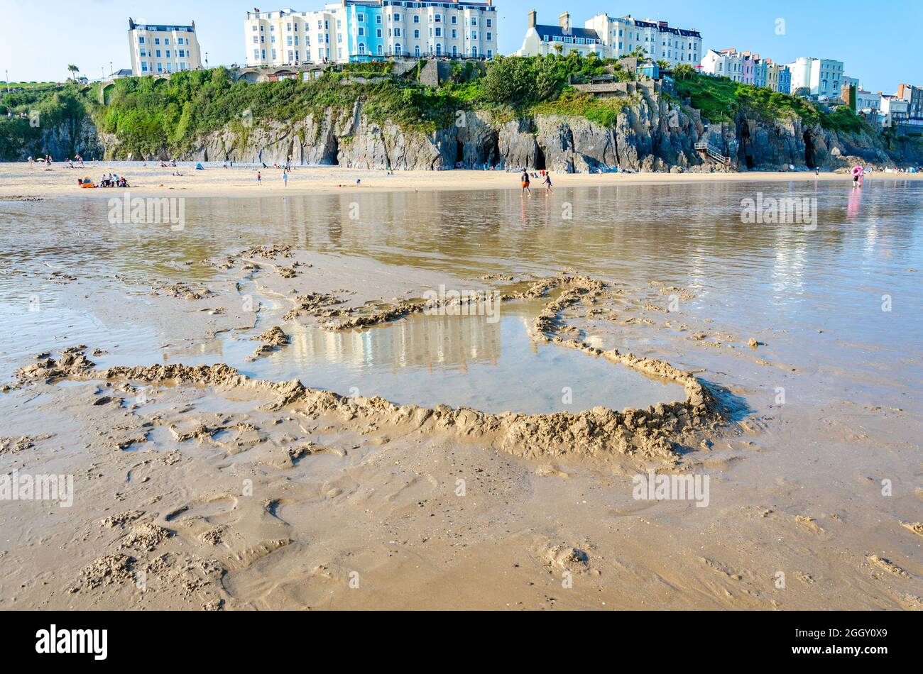 A dam built on Tenby South Beach at low tide has created a pool of water. Beyond are colourful buildings on The Esplanade ont he cliff above. Stock Photo