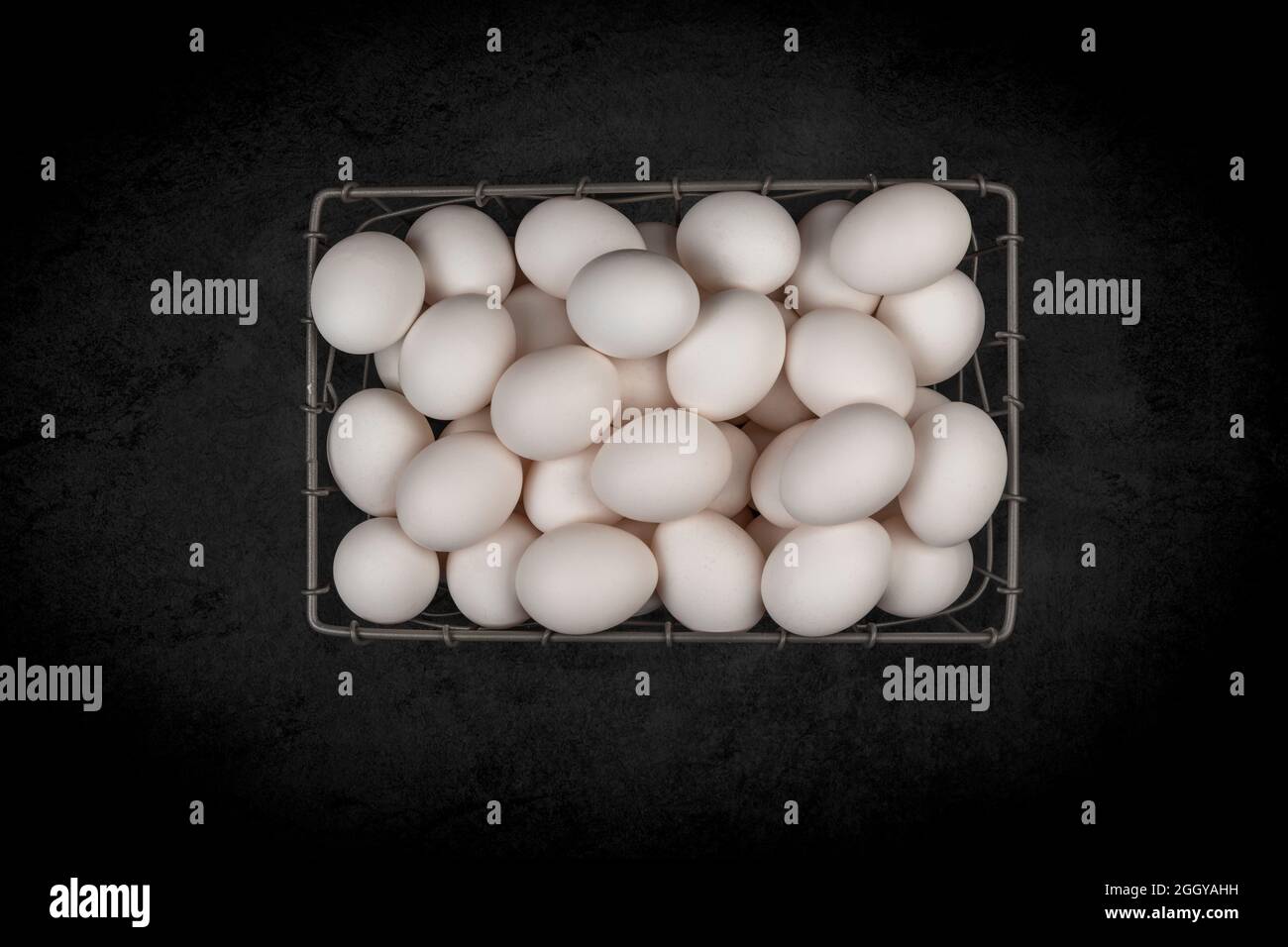 A small wire basket of assorted fresh, white eggs on a mottled black background. Stock Photo