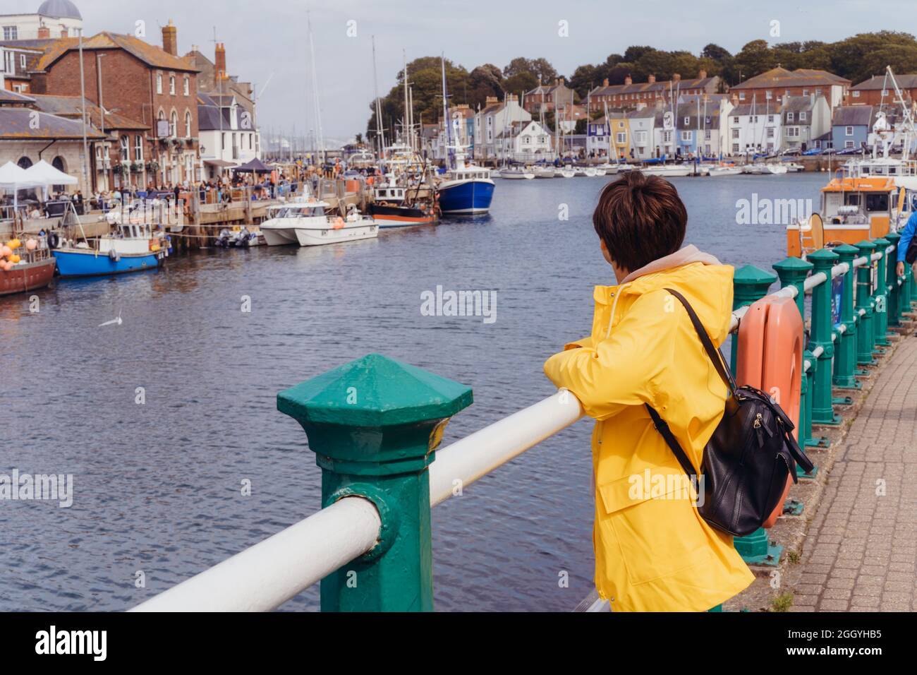 Side view woman in yellow raincoat enjoying the view of the bay with ships and boats of the seaside town. Local travel. Weymouth, England Stock Photo