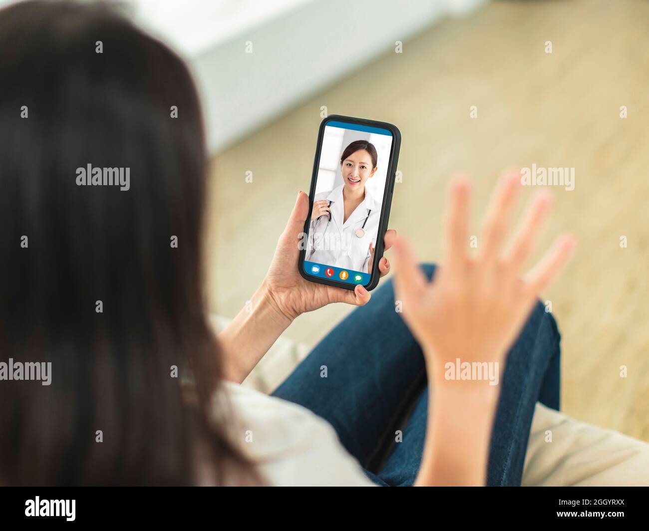 Young woman sitting on sofa at home, holding mobile phone, making video call, discussing with doctor in video conference. Stock Photo