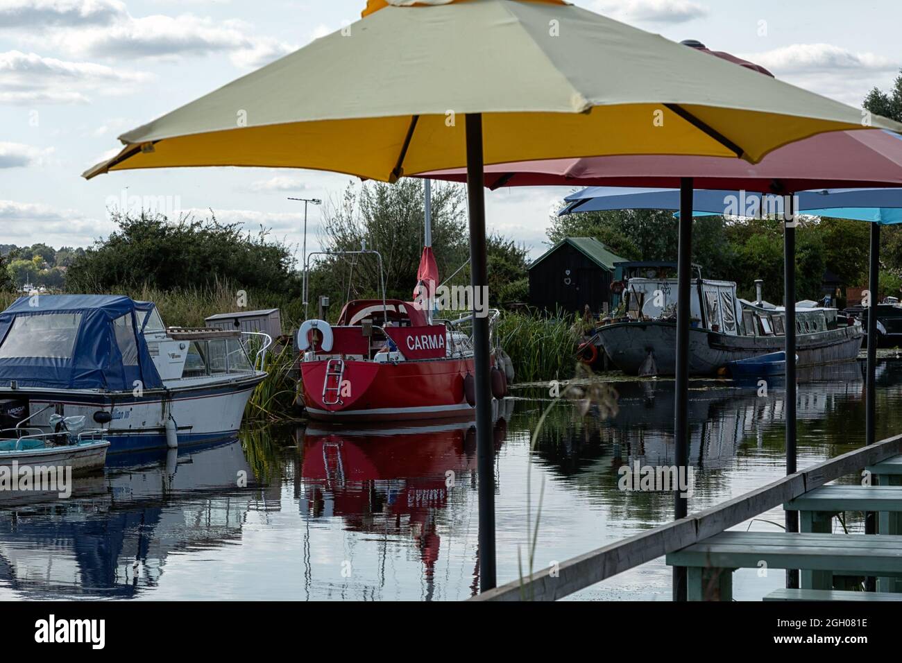 HEYBRIDGE BASIN, ESSEX, UK - AUGUST 25, 2021:  Moored boats on the Chelmer and Blackwater Navigation below umbrellas at riverside Cafe in summer Stock Photo