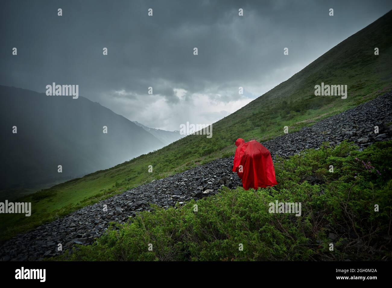 Tourist with backpack in red rain cover at beautiful scenery green mountain valley against dark rain sky in Kazakhstan Stock Photo