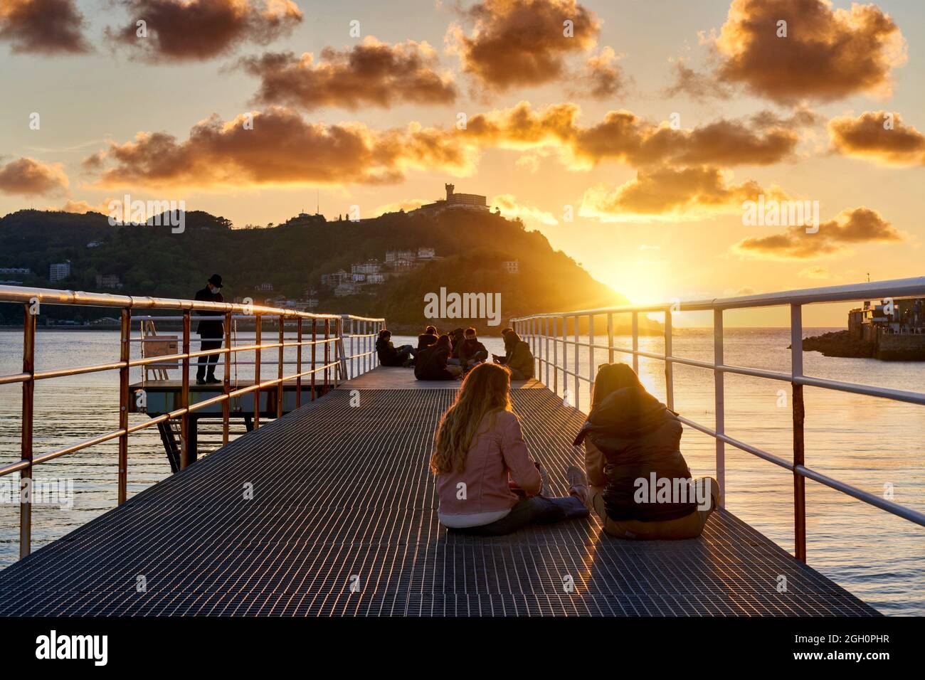 Young people enjoying the sunset at the Old Pier of the Real Club Náutico de San Sebastian, La Concha Bay, in the background Monte Igeldo, Donostia, Stock Photo