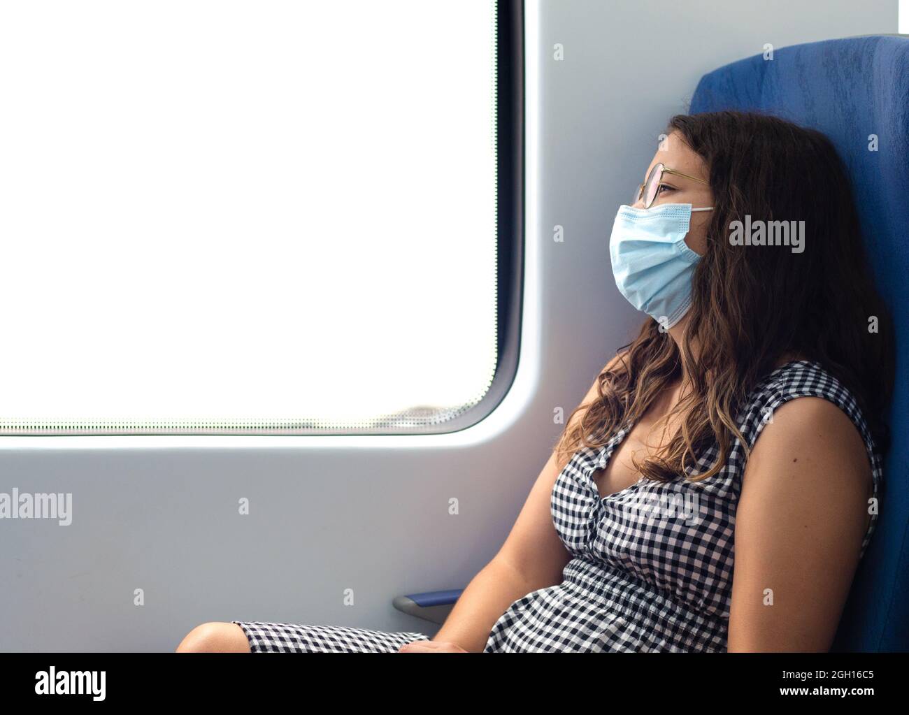 Businesswoman with protective face mask and glasses looking out of the window of train in thought. young and beautiful woman admiring the scenery whil Stock Photo