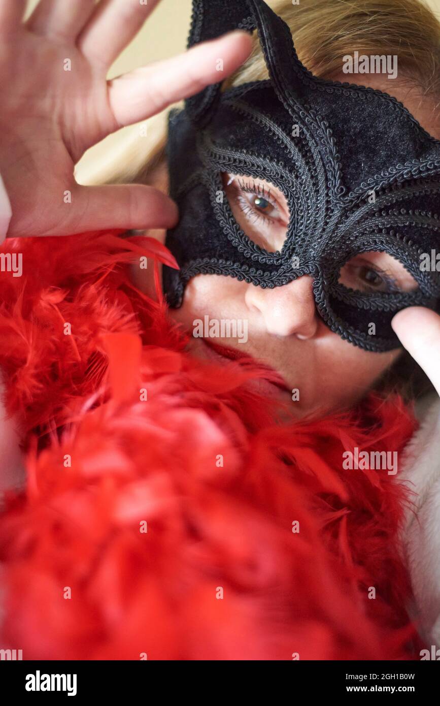 Portrait of a fifty plus years old woman with brown eyes wearing a textured black mask and red feather boa with her hand by her face. Stock Photo