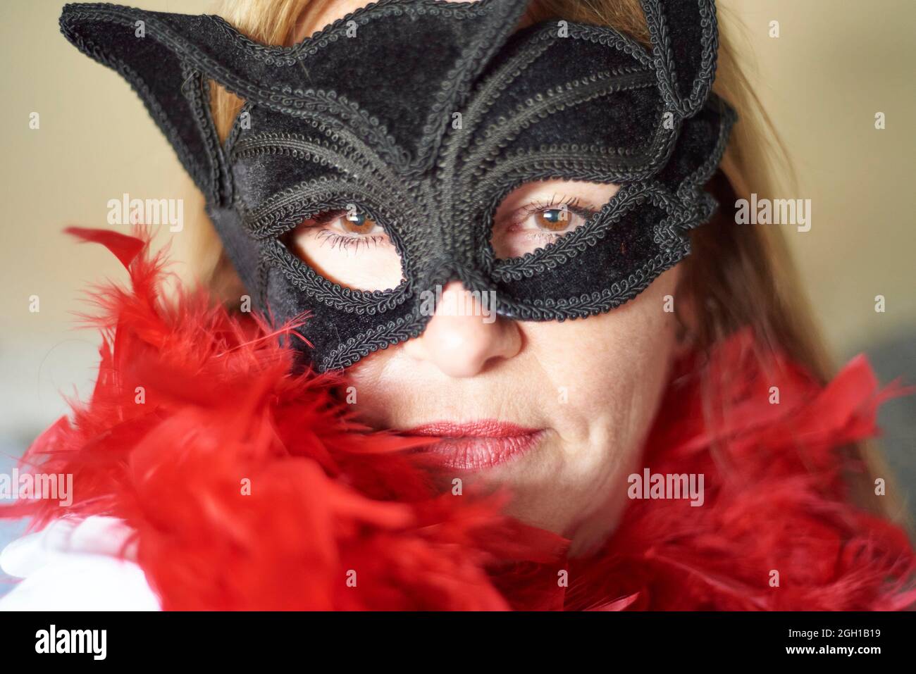 Portrait of a fifty plus years old woman with brown eyes wearing a slightly askew textured black mask and red feather boa. Stock Photo