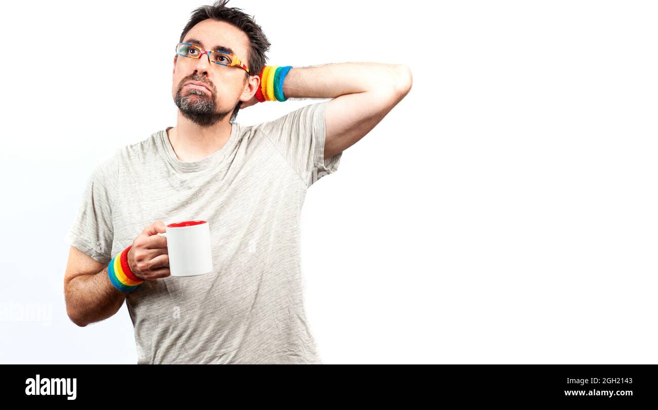 Dark haired man in gray shirt wearing glasses and a bracelet with rainbow colors; with tired expression, with one hand holding a cup . LGBTQ concept. Stock Photo