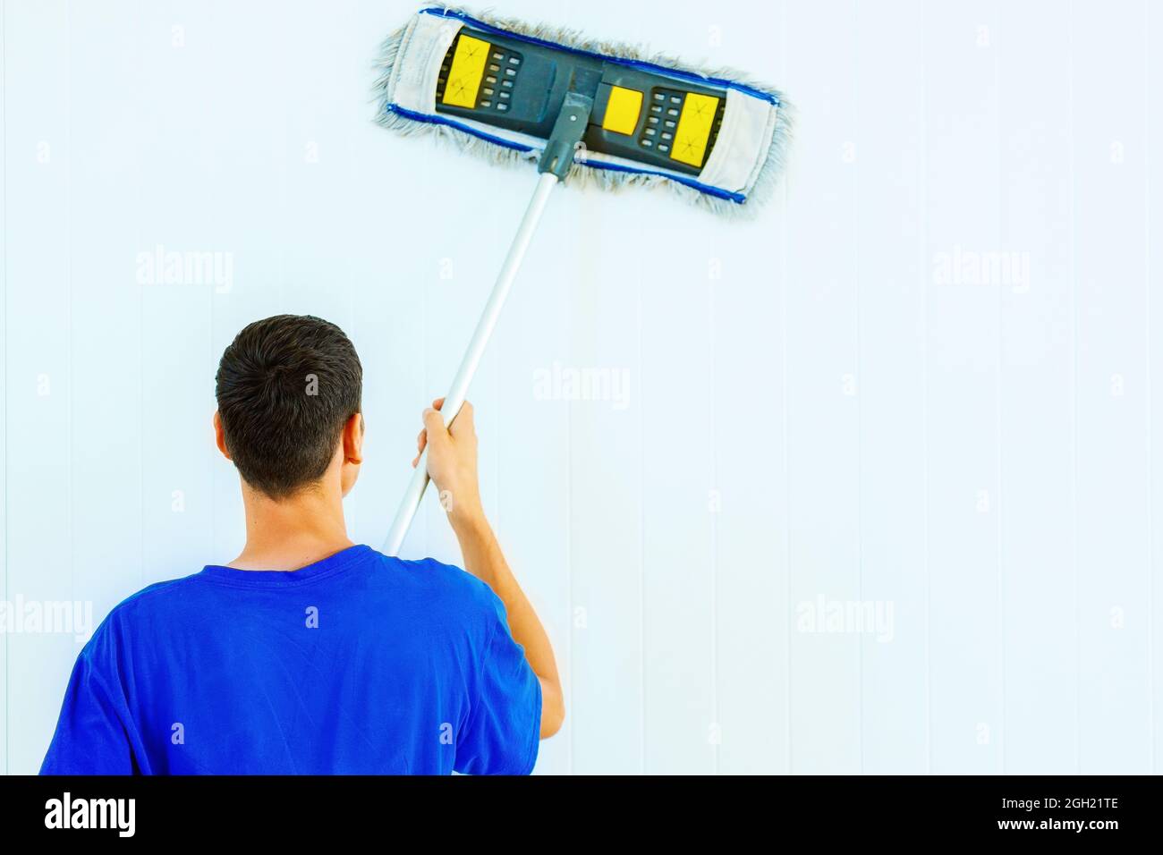 A young Caucasian man in a blue T-shirt washes the white wall of the room with a mop. Clearing service for cleaning and washing of premises. Layout Stock Photo