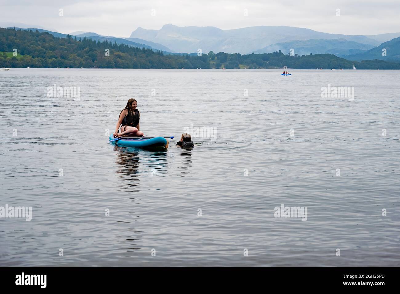 Young woman Paddle boarding with a young man swimming, in Windermere