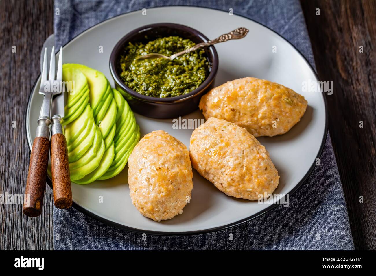 Baked in oven white fish patties or cakes served with green sauce pesto and sliced avocado on a plate on a wooden table with cutlery Stock Photo