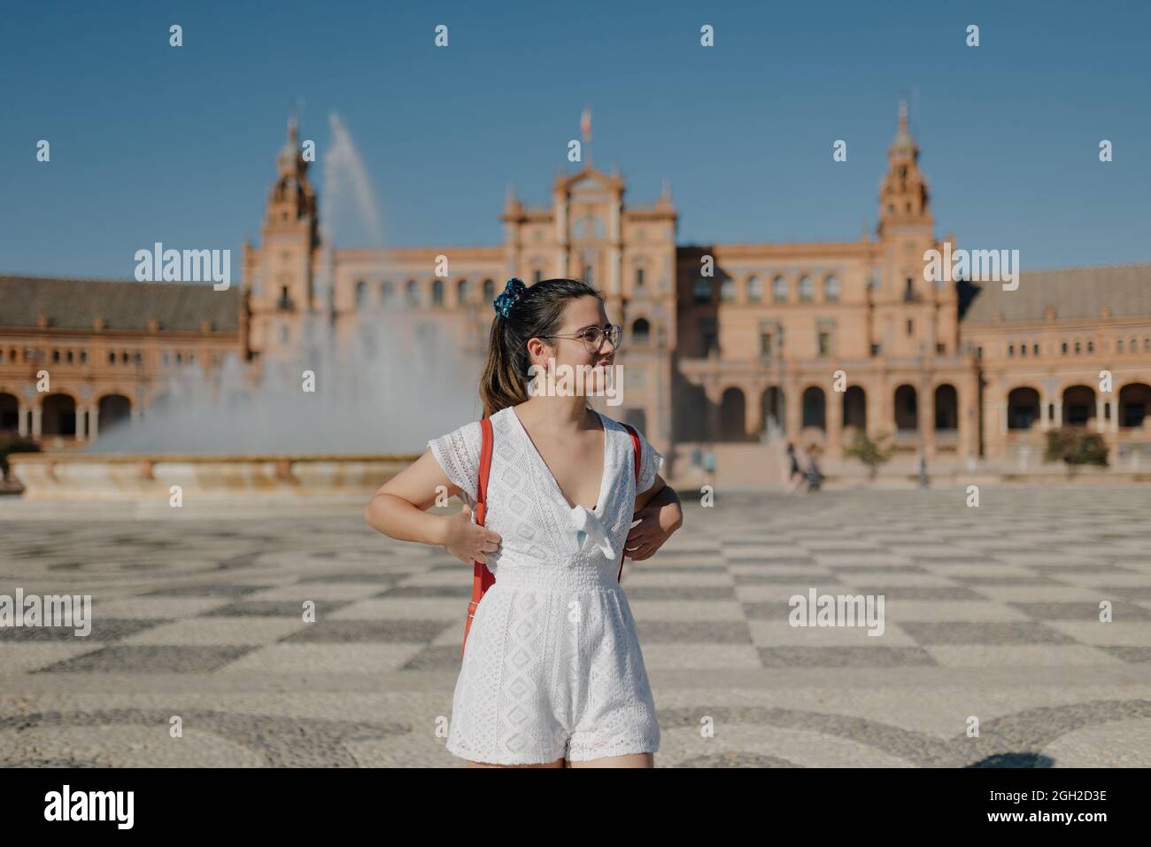 Young tourist woman with glasses wearing a white dress and red backpack is looking away and smiling while standing in the Plaza de España of Seville. Stock Photo