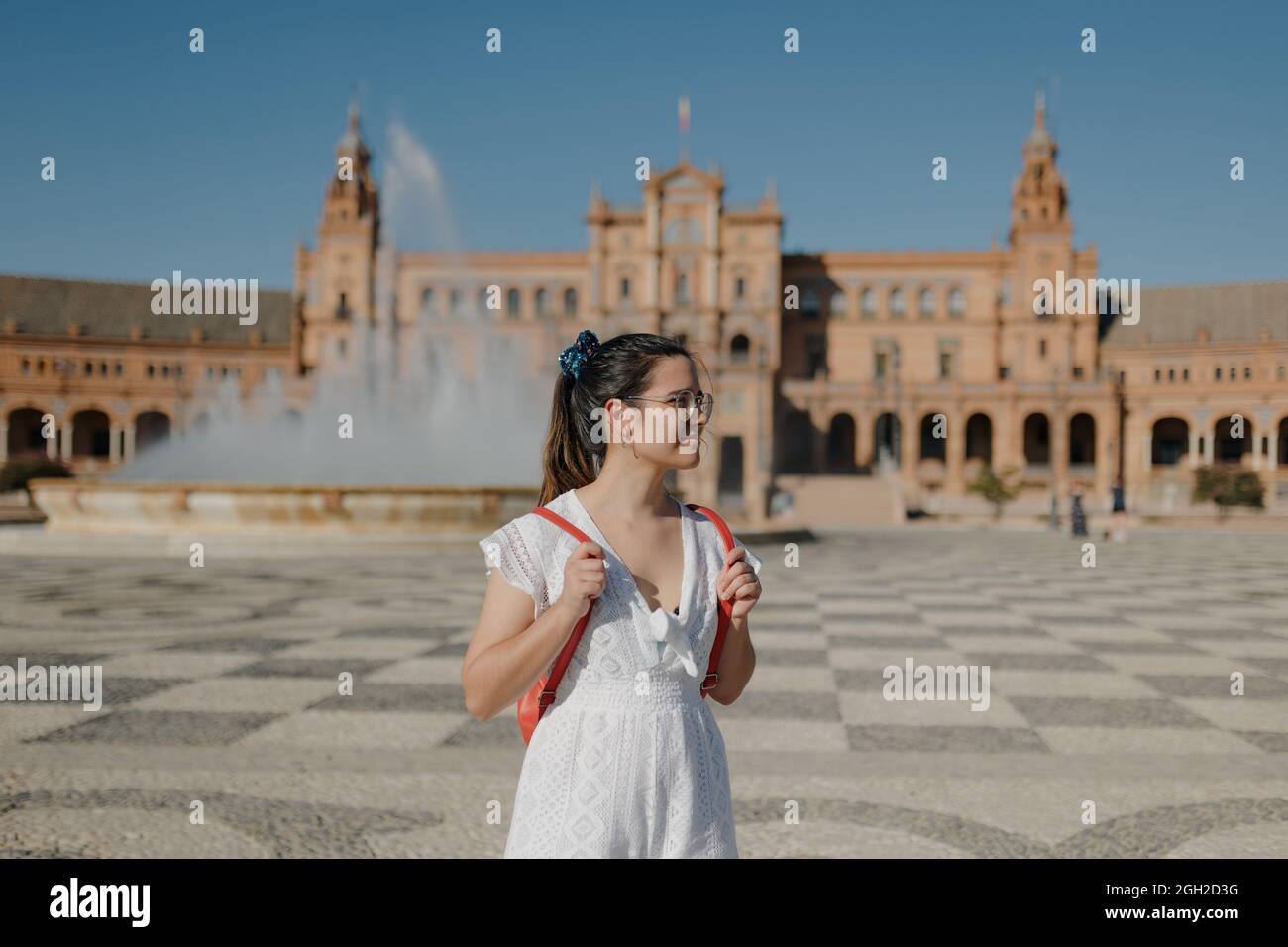 Young tourist woman with glasses wearing a white dress and red backpack is looking away and smiling while standing in the Plaza de España of Seville. Stock Photo