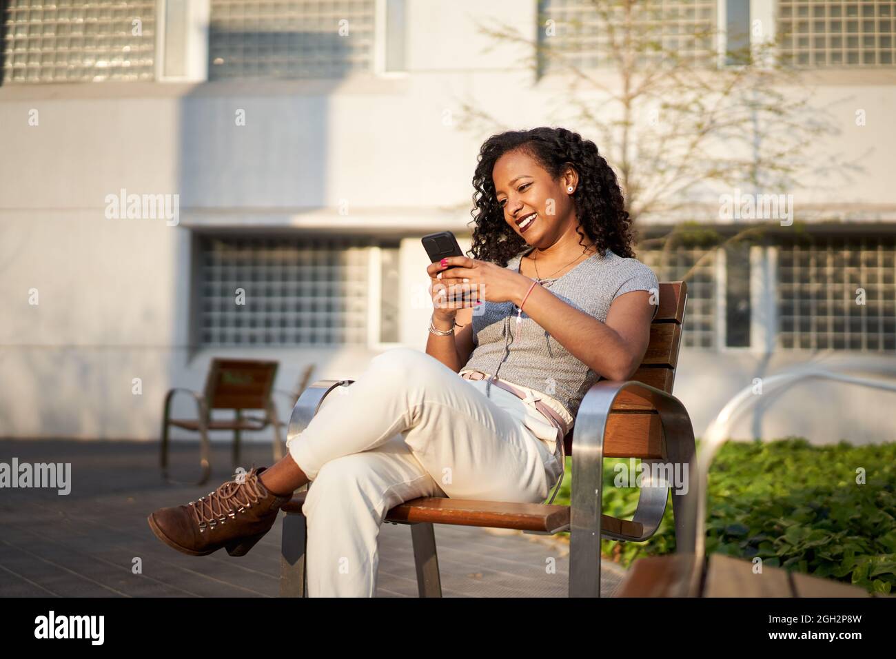 Young beautiful woman typing on cell phone sitting on bench- Smiling mixed race woman using smartphone, city at sunset.  Stock Photo