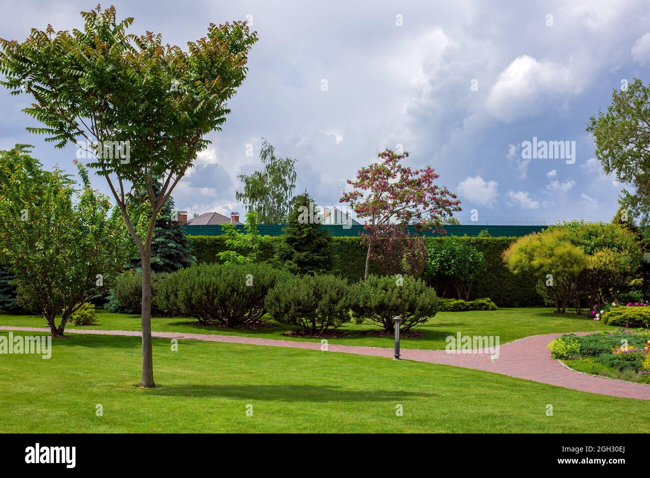 stone tile curved walkway path in park among green plants of trees with bushes and flower bed, iron ground garden lantern, nobody. Stock Photo