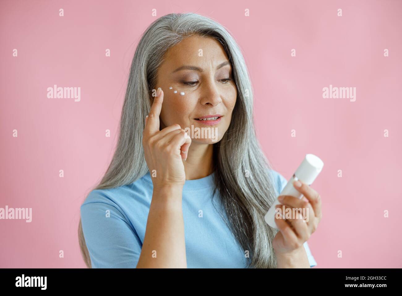Pretty middle aged Asian woman applies cream under eyes holding bottle on pink background Stock Photo