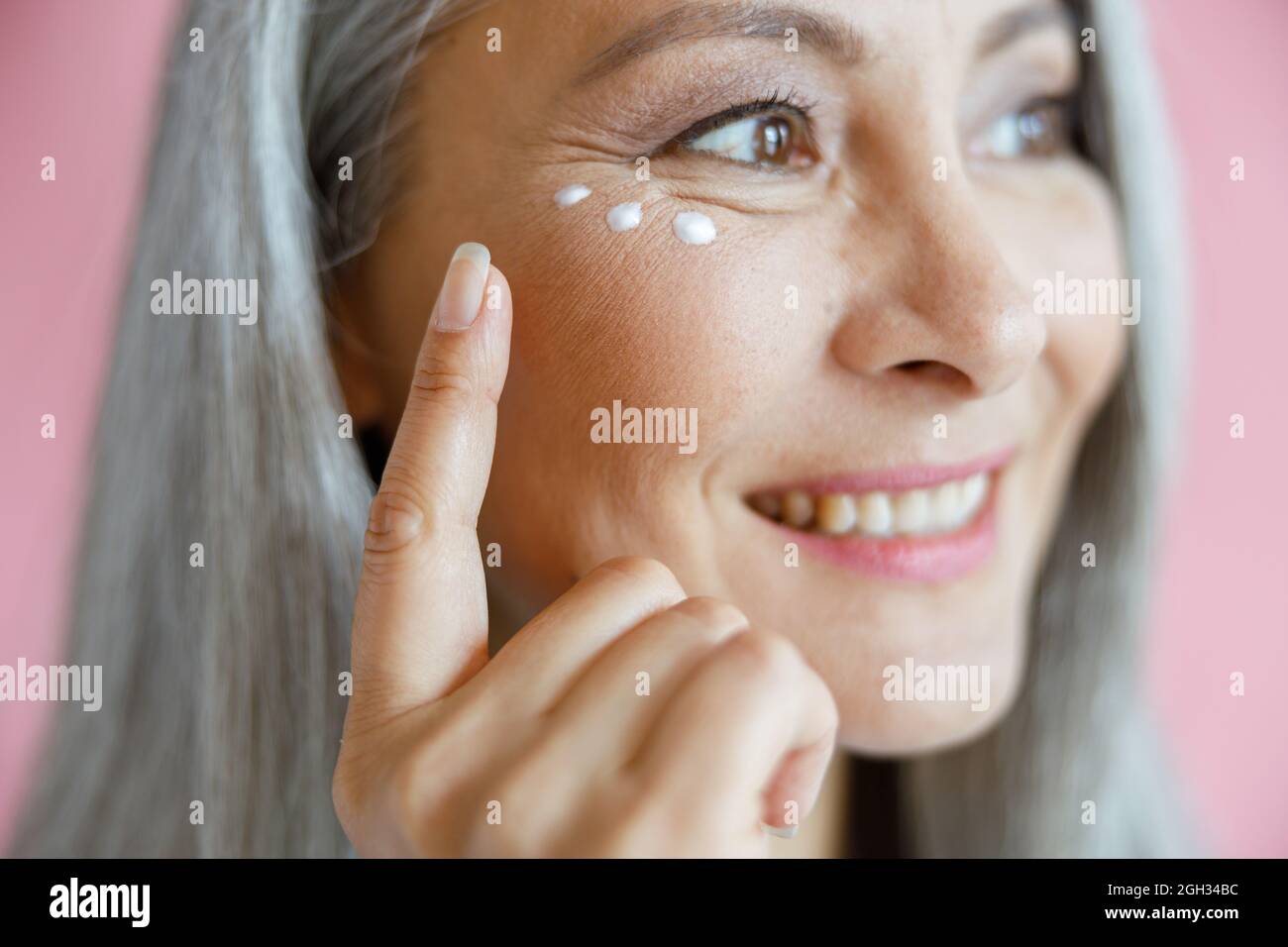 Beautiful mature Asian lady applies antiaging cream under eye on pink background Stock Photo