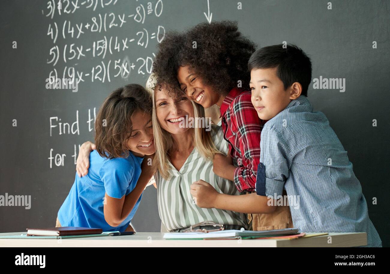 Happy diverse kids school children students hugging female teacher in classroom. Stock Photo