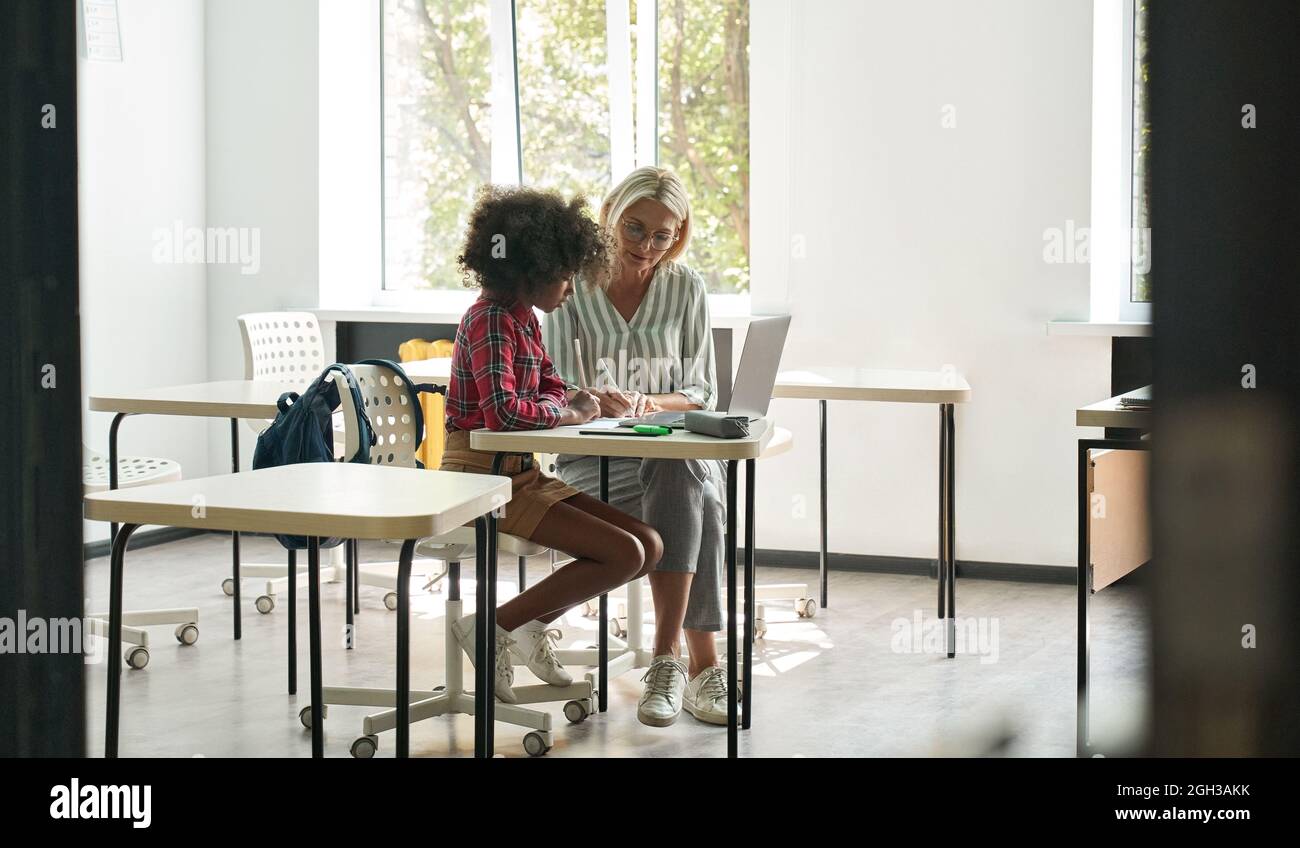 Female teacher helping African kid girl student with homework in classroom. Stock Photo