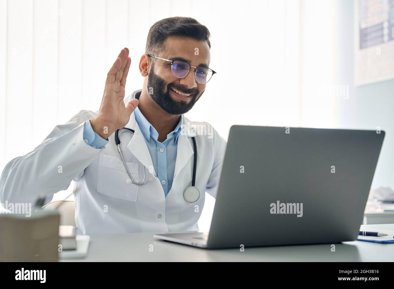 Indian male doctor waving hand having virtual online consultation on laptop. Stock Photo