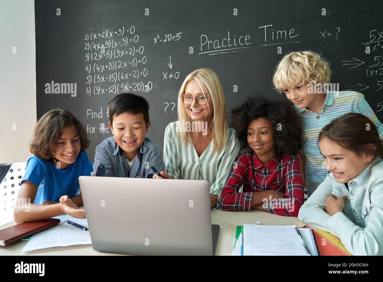 Happy diverse school children students gather at teacher table look at laptop. Stock Photo