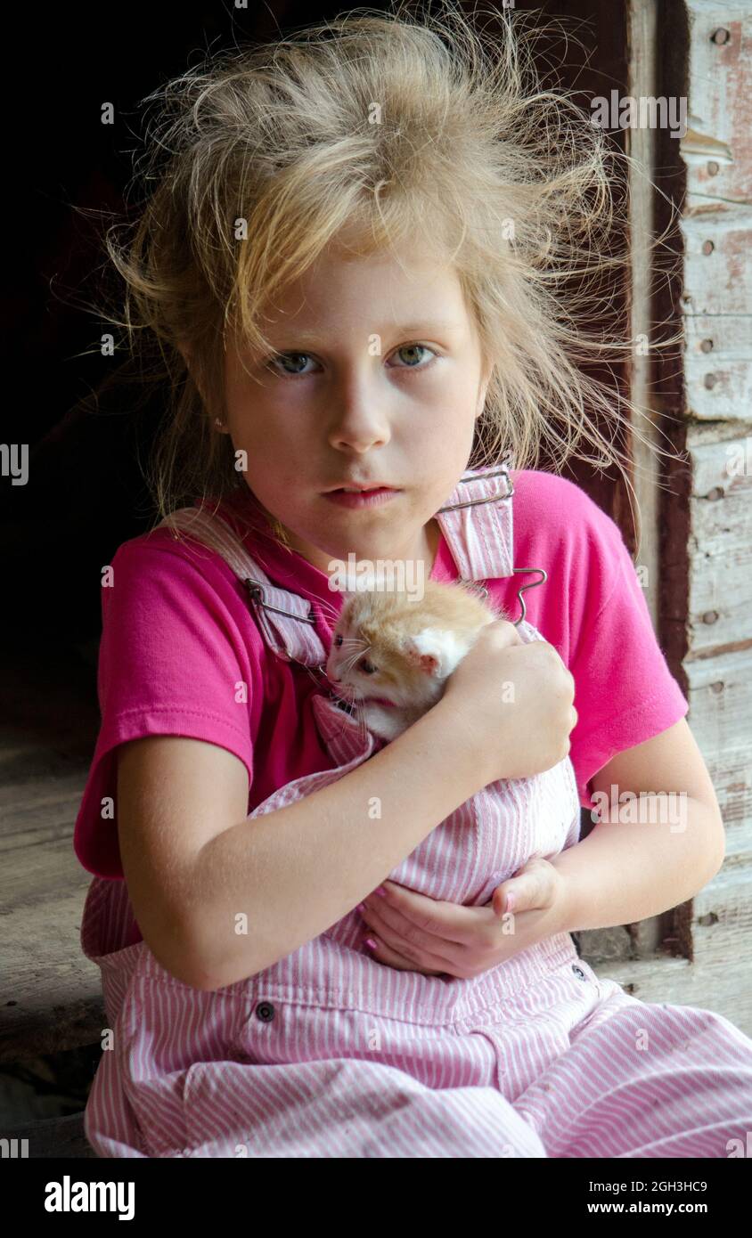 An adorable little farm girl in pink overalls hugs a small kitten to her chest as she sits on a step outside the barn Stock Photo