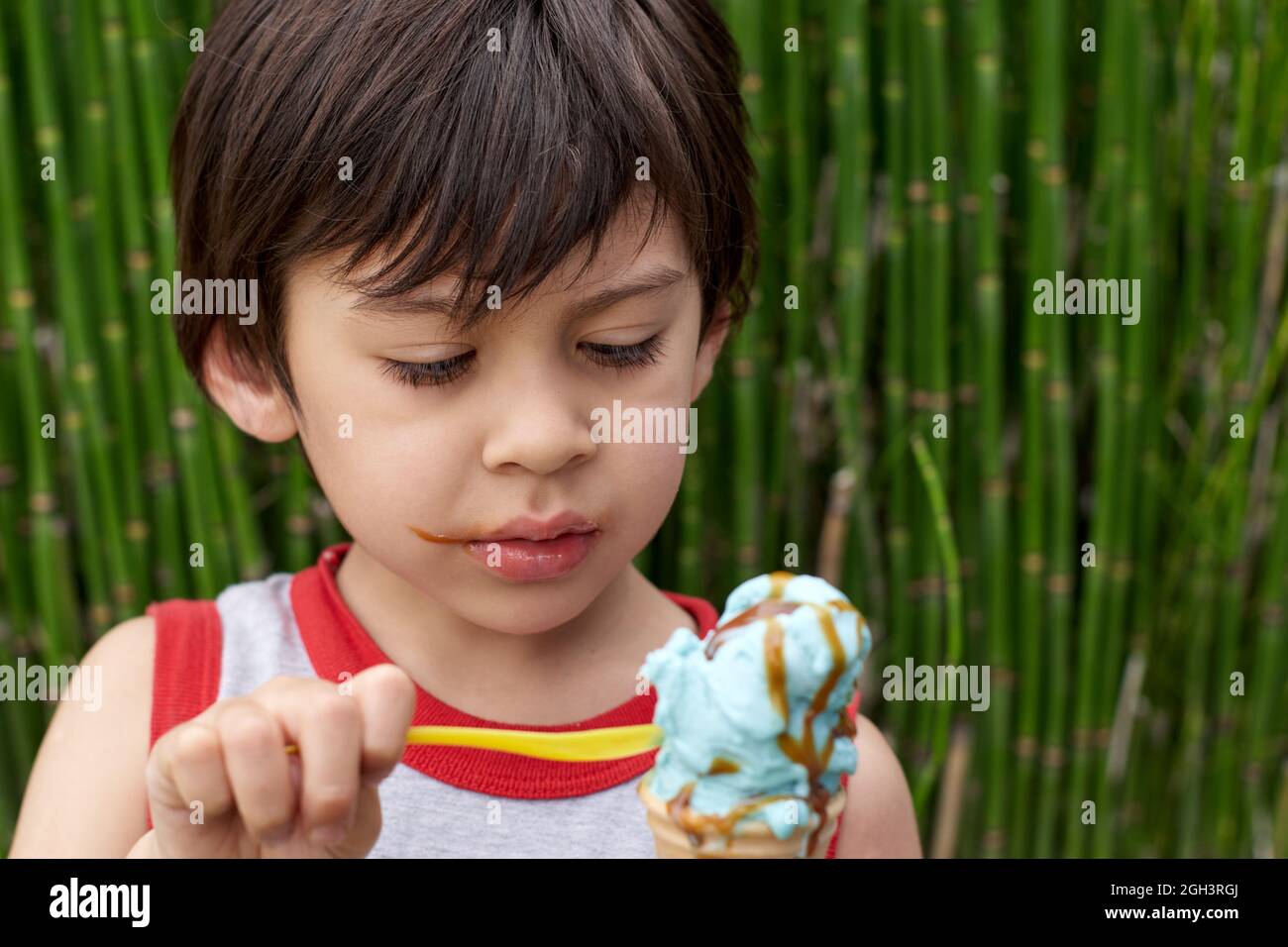 Brunette little boy eating ice cream cone with a spoon wearing sleeveless shirt in summertime. Green plants on background. Horizontal Stock Photo