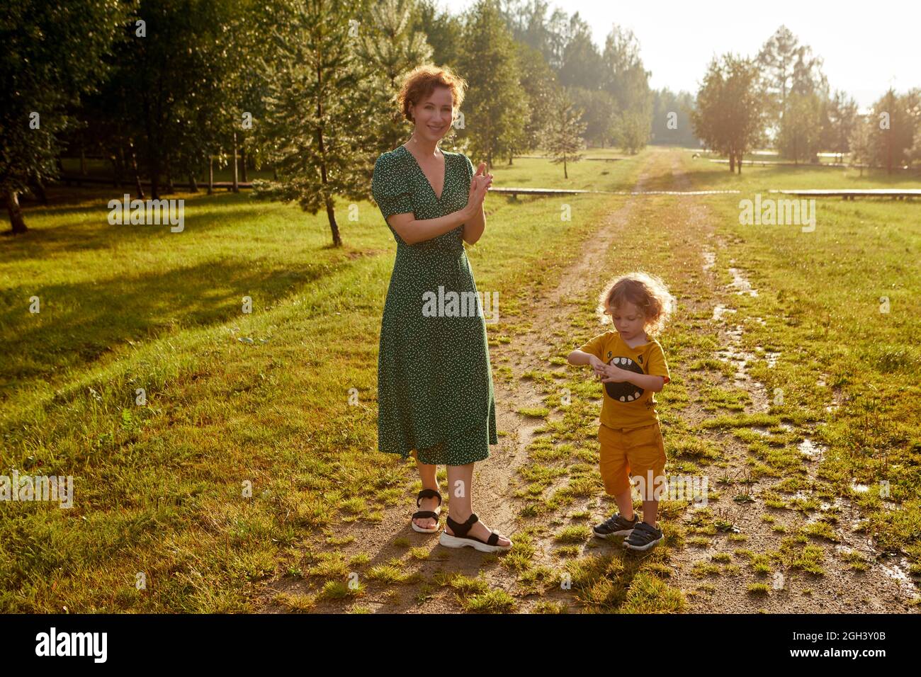 Full body of positive mother looking at camera while standing on grassy pathway with cute son near trees in nature Stock Photo