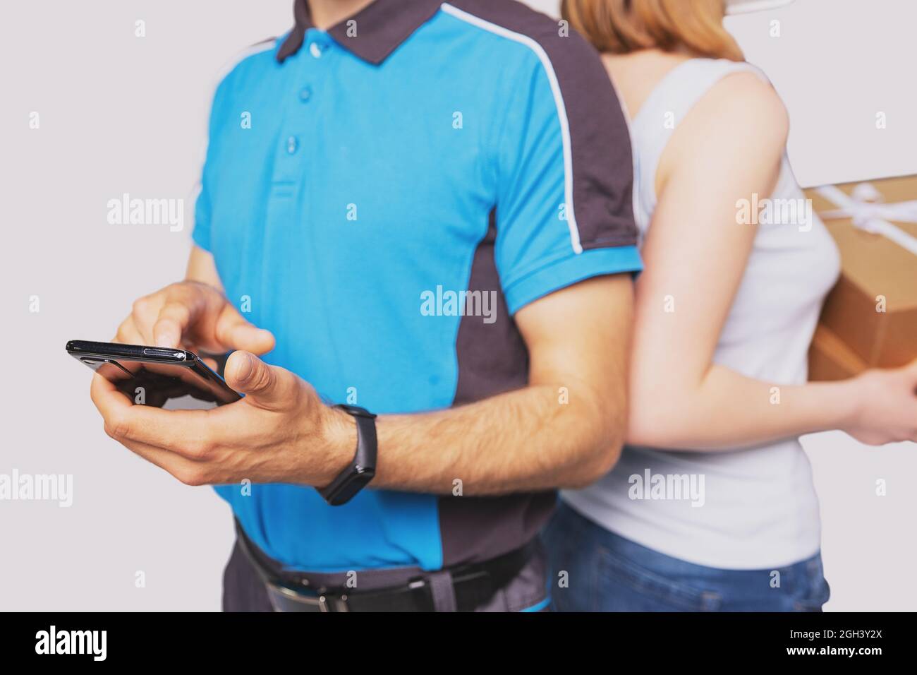 Cropped photo of a postman and a woman blonde standing back to back, postman touching the mobile phone in his hand and woman holding the package. Stock Photo