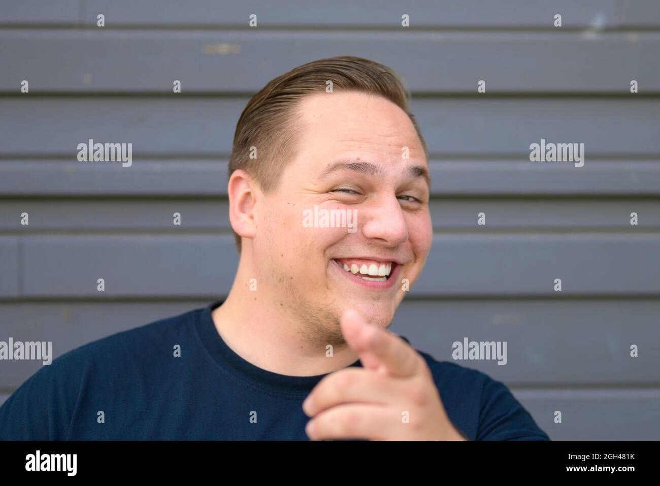 Gleeful young man pointing at the camera with a beaming smile of amusement as he poses against a grey exterior wall with focus to his face Stock Photo