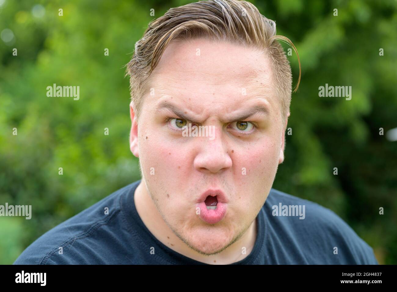 Angry aggressive young man shouting at the camera with a threatening expression in a close up on his face outdoors against greenery Stock Photo